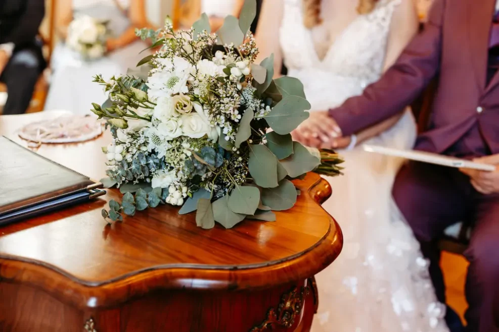 Nahaufnahme eines Straußes aus weißen Blumen und Grünzeug auf einem polierten Holztisch, mit einer Braut in einem weißen Kleid und einem Bräutigam in einem violetten Anzug, die zusammensitzen und sich an den Händen halten, im Hintergrund.
