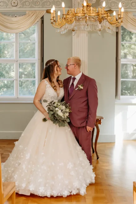 Eine Braut in einem weißen Blumenkleid und ein Bräutigam in einem lilafarbenen Anzug stehen sich lächelnd in einem eleganten Raum mit großen Fenstern und einem Kronleuchter gegenüber. Die Braut hält einen Strauß aus grünen und weißen Blumen in der Hand.