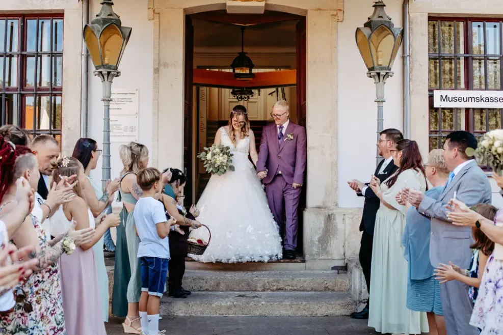 Eine Braut in einem weißen Kleid und ein Mann in einem violetten Anzug treten lächelnd aus einem Gebäude, während eine Menschenmenge auf beiden Seiten klatscht und feiert. Die Szenerie wirkt festlich und fröhlich.