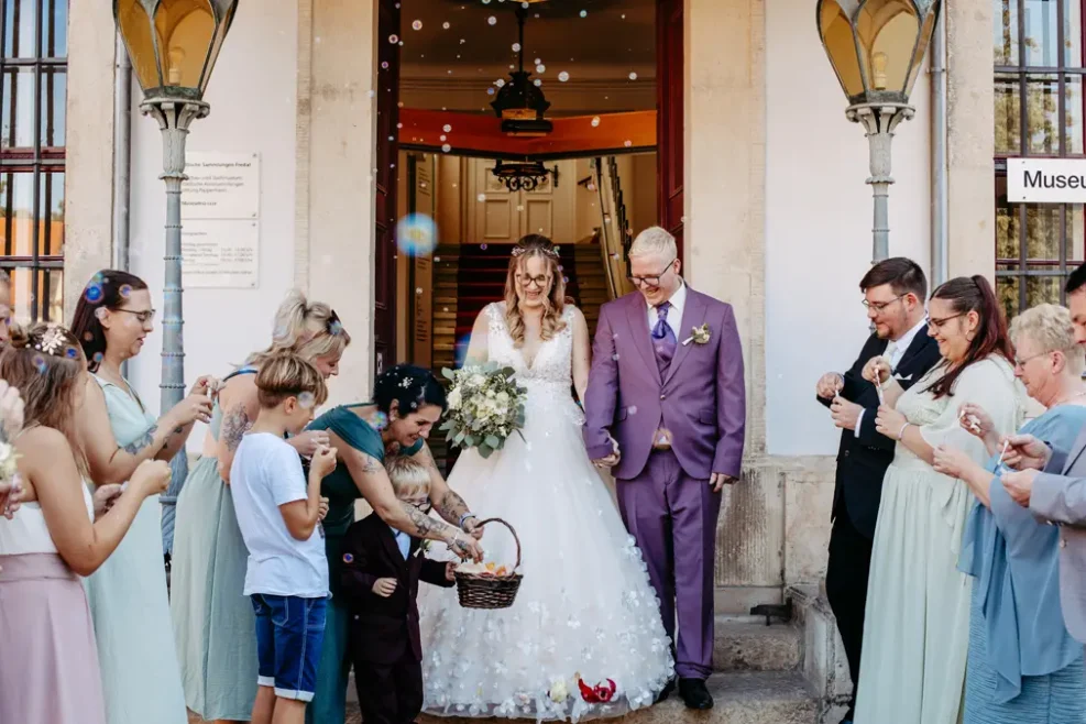 Eine Braut und ein Bräutigam, umgeben von lächelnden Gästen, stehen auf einer Treppe vor einem Gebäude. Die Braut trägt ein weißes Hochzeitskleid, der Bräutigam einen lavendelfarbenen Anzug, und Seifenblasen schweben, während ein Blumenmädchen Blütenblätter aus einem Korb verstreut.