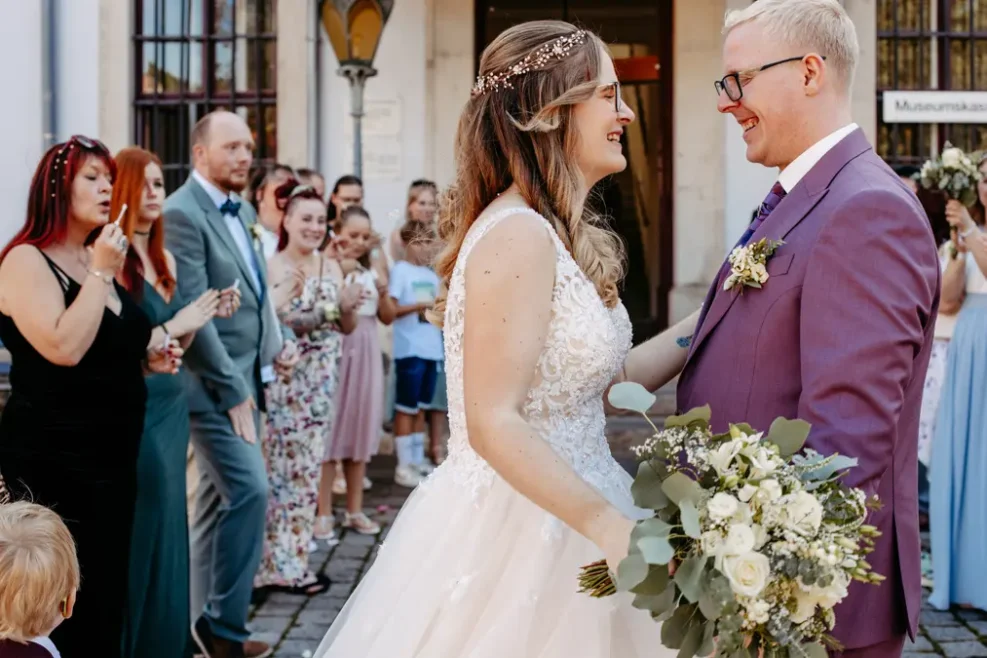 Eine Braut und ein Bräutigam lächeln sich an, während sie sich an den Händen halten, umgeben von jubelnden Freunden und Familienmitgliedern bei einer Hochzeit im Freien. Die Braut trägt ein weißes Kleid und hält einen Blumenstrauß, der Bräutigam trägt einen lilafarbenen Anzug.