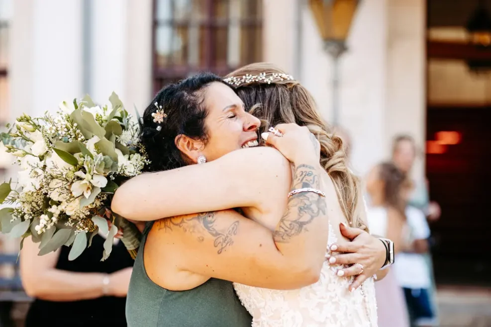 Zwei Frauen umarmen sich und lächeln während einer Hochzeitsfeier; die eine hält einen Strauß weißer Blumen, die andere trägt ein Spitzenkleid und ein juwelenbesetztes Haarteil. Der Hintergrund ist leicht unscharf, Menschen und Architektur sind zu erkennen.