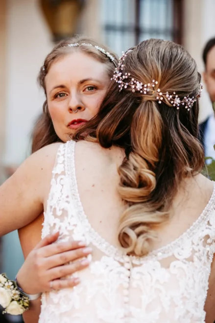Zwei Bräute in Hochzeitskleidern umarmen sich eng. Die eine Braut, die der Kamera zugewandt ist, hat langes helles Haar und einen mit Juwelen besetzten Kopfschmuck; die andere steht mit dem Rücken zur Kamera und zeigt detaillierte Spitzen und Perlen an ihrem Kleid.