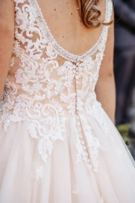Nahaufnahme einer Frau, die ein Hochzeitskleid mit aufwändiger Spitzenverzierung und Perlenknöpfen am Rücken trägt. Das Kleid hat einen tiefen Rückenausschnitt und zarte Stickereien.