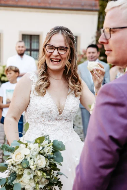 Eine lächelnde Braut in einem Spitzenhochzeitskleid hält einen Strauß weißer Blumen, umgeben von Gästen im Freien. Ein Mann mit Brille steht im Vordergrund, und im Hintergrund sind Menschen versammelt.
