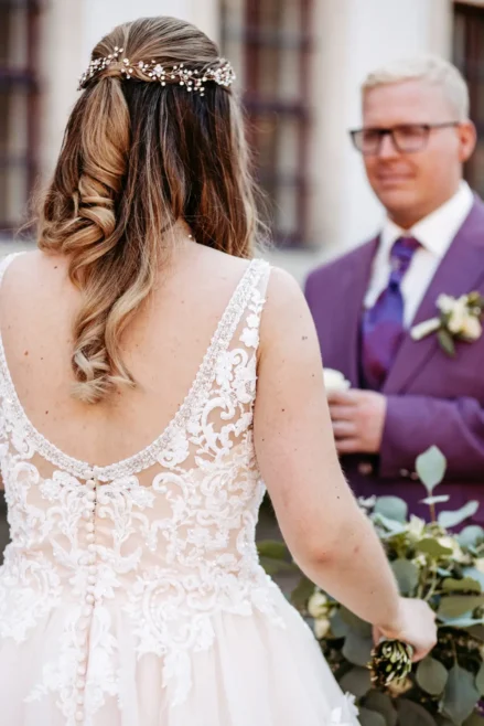 Eine Braut in einem Spitzenhochzeitskleid steht mit dem Rücken zur Kamera vor einem Bräutigam in einem lila Anzug, einer Brille und einer Ansteckblume in der Hand, während einer Hochzeitszeremonie im Freien.