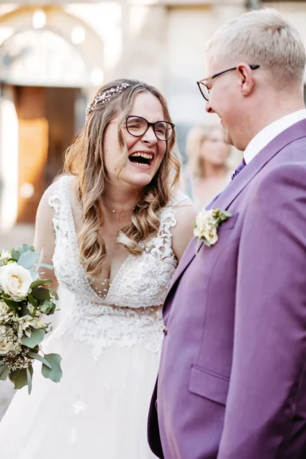 Eine Braut in einem weißen Spitzenhochzeitskleid mit Brille lacht fröhlich, während sie neben einem Bräutigam in einem lavendelfarbenen Anzug mit Brille steht. Sie hält einen Strauß weißer Blumen in der Hand, und die beiden sehen sich glücklich an.