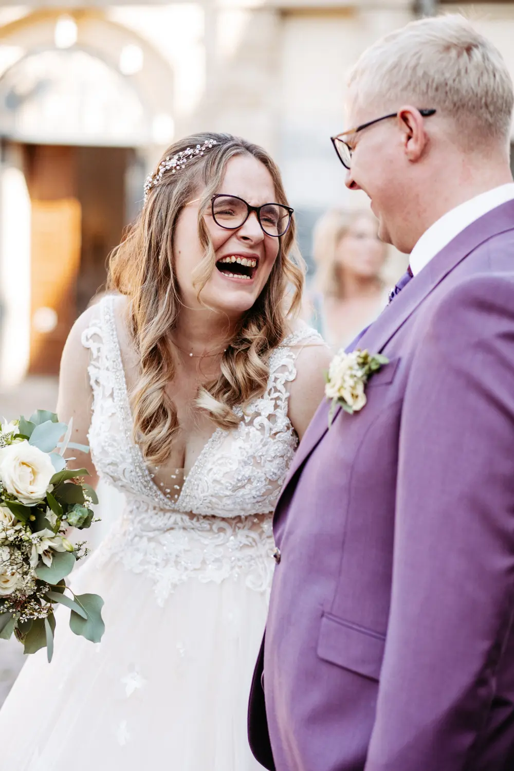 Eine Braut in einem weißen Spitzenhochzeitskleid mit Brille lacht fröhlich, während sie neben einem Bräutigam in einem lavendelfarbenen Anzug mit Brille steht. Sie hält einen Strauß weißer Blumen in der Hand, und die beiden sehen sich glücklich an.