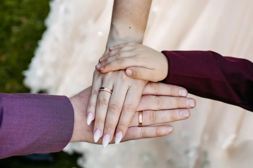Drei Hände - zwei Erwachsene mit Eheringen und ein Kind - sind übereinandergelegt und symbolisieren die Einheit. Die Erwachsenen tragen passende violette Ärmel, und im Hintergrund ist ein weißes Kleid zu sehen.