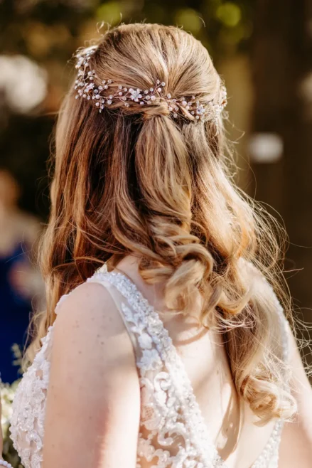 Eine Frau mit gewelltem blondem Haar, das halb hochgesteckt und mit einem zarten Blumenhaarteil geschmückt ist, trägt ein weißes Spitzenhochzeitskleid und wird von hinten in einer weichen Außenbeleuchtung gezeigt.