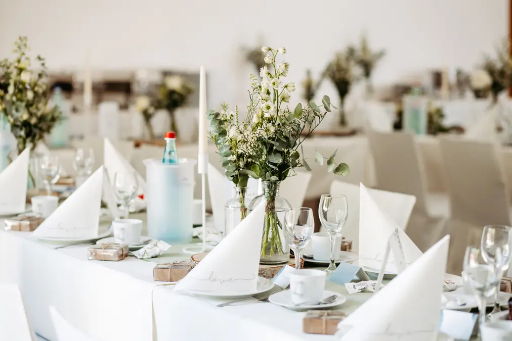 Ein langer, elegant gedeckter Tisch mit weißen Servietten, Gläsern, Teetassen und grünen Blumen in der Mitte; eine Flasche Wasser steht in einem gefrosteten Halter. Das Dekor ist hell und minimalistisch und eignet sich für eine formelle Veranstaltung oder eine Hochzeit.
