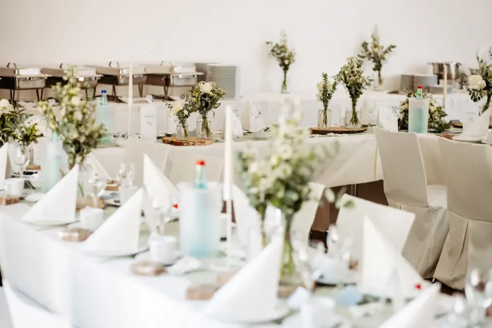 Elegante Banketttische mit weißen Tischtüchern, weißen Servietten, Gläsern, Tellern und Blumenschmuck. Der Hintergrund zeigt weitere Tische, Blumen und abgedeckte Chafing Dishes.
