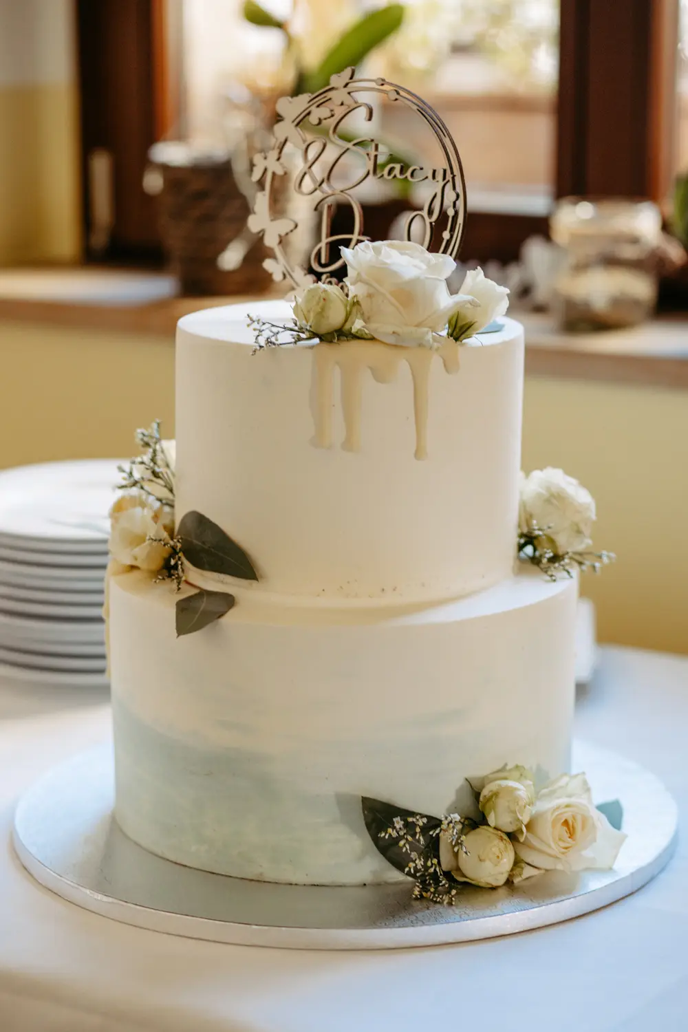 Eine zweistöckige weiße Hochzeitstorte, dekoriert mit weißen Rosen, Grünzeug und einem personalisierten Topper. Die Torte hat einen subtilen blauen Aquarell-Effekt auf der unteren Etage und weiße Schokoladentropfen auf der oberen Etage. Die Teller sind in der Nähe gestapelt.