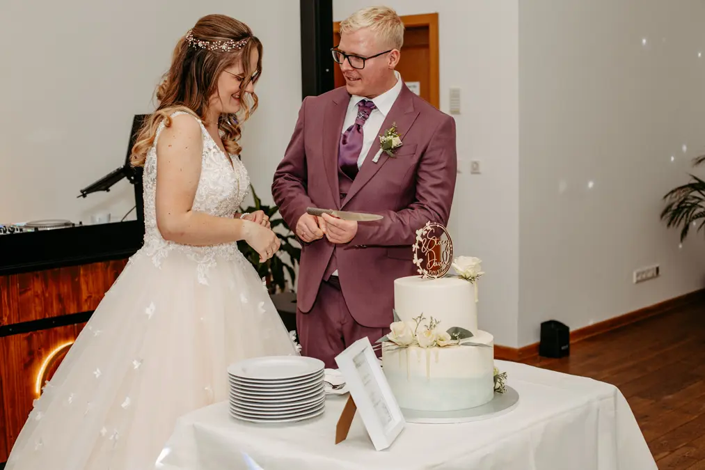 Eine Braut und ein Bräutigam stehen neben einer dekorierten Hochzeitstorte und bereiten sich lächelnd auf das Anschneiden der Torte vor. Die Braut trägt ein weißes Kleid, der Bräutigam einen lilafarbenen Anzug mit einer lila Krawatte. Auf dem Tisch vor ihnen sind Teller gestapelt.