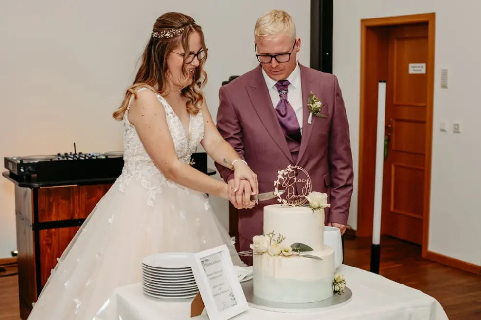 Eine Braut und ein Bräutigam, beide mit Brille, schneiden gemeinsam eine weiße Hochzeitstorte an. Die Braut trägt ein weißes Kleid und der Bräutigam einen malvenfarbenen Anzug. Auf dem Tisch neben der Torte stehen Teller und ein gerahmtes Schild.