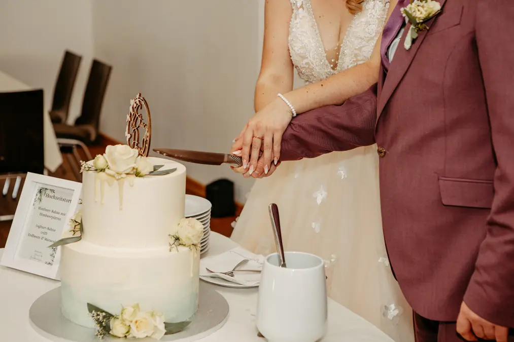 Eine Braut und ein Bräutigam schneiden gemeinsam eine zweistöckige weiße Hochzeitstorte an, wobei sie das Messer in der Hand halten. Die Torte ist mit weißen Blumen und einem Topper geschmückt, und in der Nähe steht eine Vase mit Utensilien. Ihre Gesichter sind nicht zu sehen.