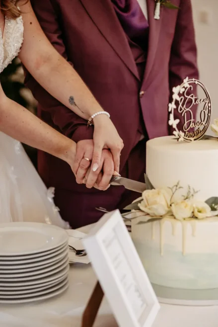 Eine Braut und ein Bräutigam schneiden gemeinsam eine weiße Hochzeitstorte an, wobei sich ihre Hände auf dem Messer überlappen. Die Torte ist mit weißen Rosen und einem individuellen Topper verziert. Auf dem Tisch sind gestapelte Teller und ein gerahmtes Schild zu sehen.