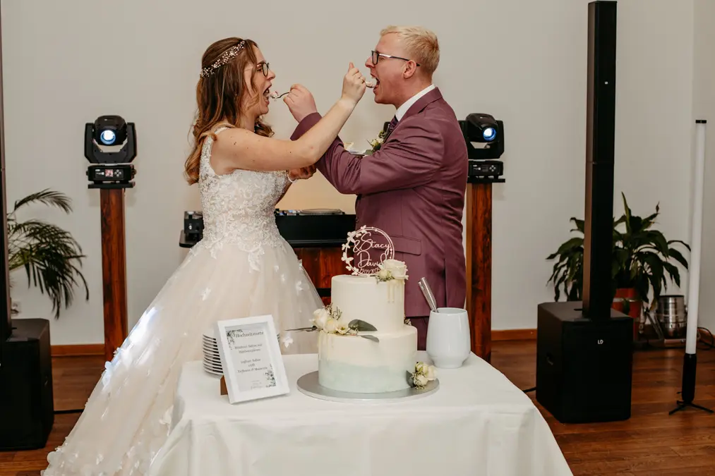 Eine Braut und ein Bräutigam lächeln und füttern sich gegenseitig mit Kuchen auf ihrer Hochzeitsfeier. Sie stehen neben einer weißen Stufentorte auf einem dekorierten Tisch in einer Halle.