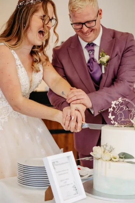 Eine fröhliche Braut und ein Bräutigam, beide mit Brille, lachen beim Anschneiden ihrer weißen Hochzeitstorte. Der Bräutigam trägt einen lila Anzug, die Braut ein Spitzenkleid. Ein Tortenaufleger und eine gerahmte Speisekarte stehen auf dem Tisch.