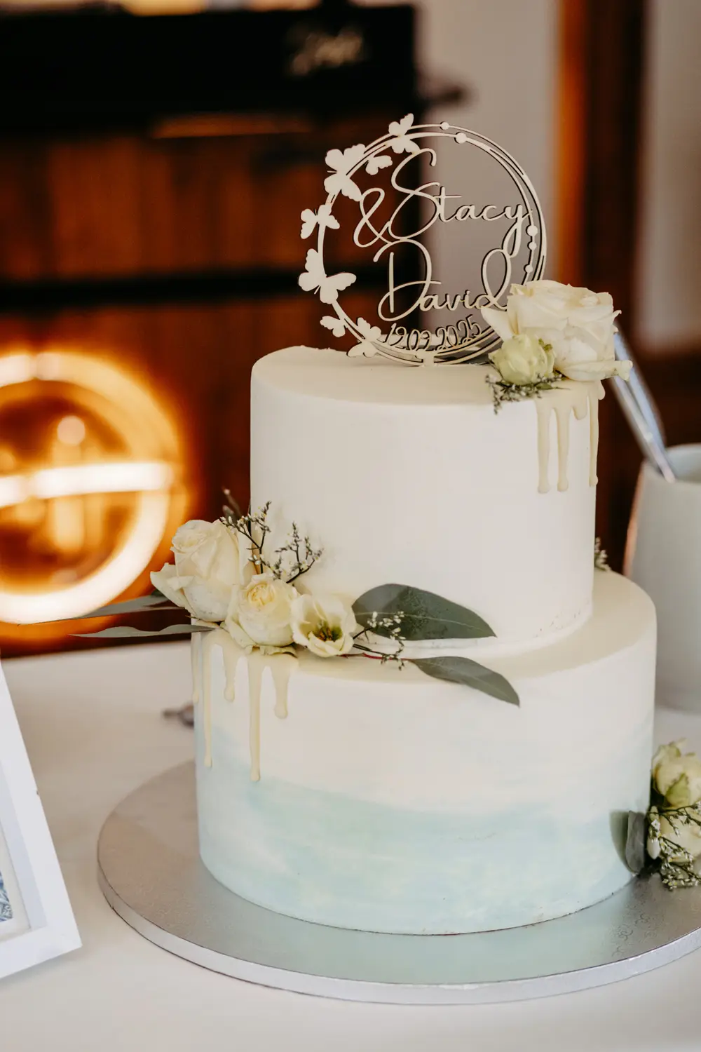 Eine zweistöckige weiße Hochzeitstorte mit blassblauen Akzenten, dekoriert mit weißen Rosen und Grünzeug, weißen Schokoladentropfen und einem Topper mit der Aufschrift Stacy & David. Die Torte steht auf einem runden Silberbrett.