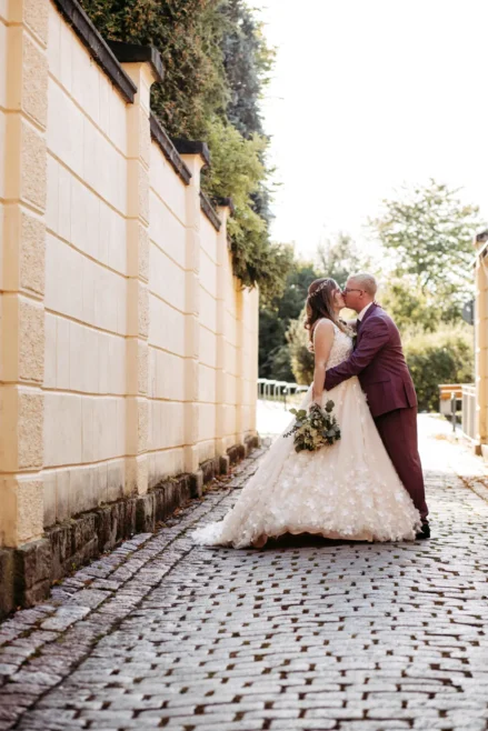 Eine Braut und ein Bräutigam stehen in einer Kopfsteinpflaster-Gasse, umarmen sich und küssen sich. Die Braut trägt ein langes, weißes Kleid und hält einen Blumenstrauß in der Hand, während der Bräutigam einen violetten Anzug trägt. Bäume und eine sonnenbeschienene Mauer säumen den Weg.