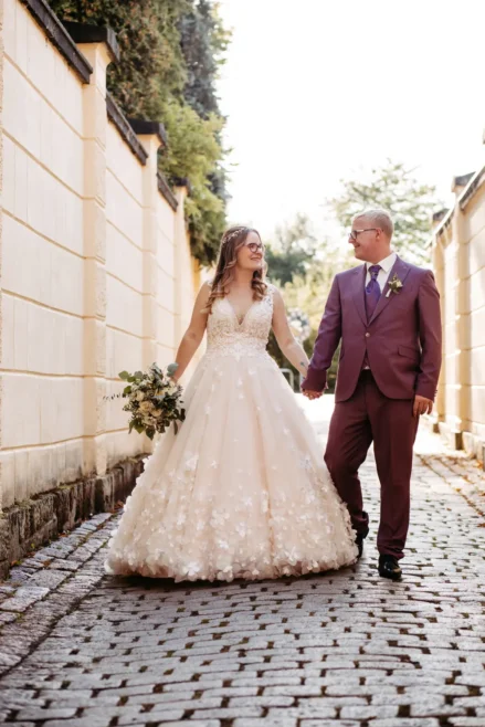 Eine Braut in einem weißen, blumengeschmückten Kleid und ein Bräutigam in einem violetten Anzug gehen an einem sonnigen Tag Hand in Hand einen kopfsteingepflasterten Weg entlang und lächeln sich an.