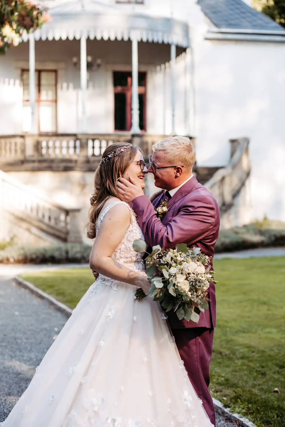 Eine Braut in einem weißen Kleid und ein Bräutigam in einem lilafarbenen Anzug lächeln sich im Freien liebevoll an, wobei der Bräutigam einen Blumenstrauß hält und das Gesicht der Braut sanft berührt; im Hintergrund ist ein weißes Gebäude zu sehen.