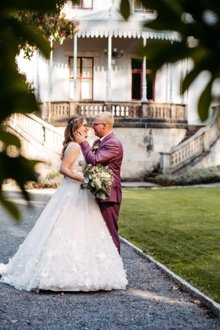 Eine Braut und ein Bräutigam stehen im Freien zusammen und lächeln sich an, wobei der Bräutigam das Gesicht der Braut berührt. Die Braut trägt ein weißes Kleid und hat einen Blumenstrauß in der Hand; der Bräutigam trägt einen lilafarbenen Anzug. Im Hintergrund ist ein historisches Gebäude zu sehen.