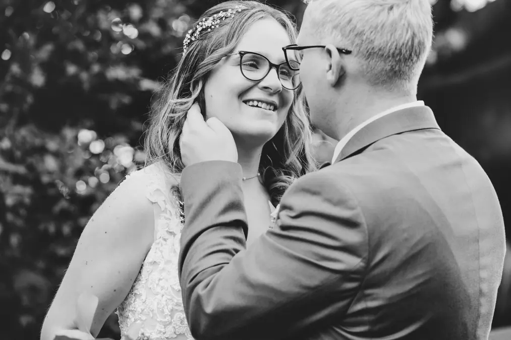 Ein Schwarz-Weiß-Foto einer lächelnden Braut und eines lächelnden Bräutigams, die sich gegenüberstehen. Der Bräutigam berührt sanft das Gesicht der Braut, und beide tragen eine Brille. Der Hintergrund ist unscharf mit Grünpflanzen im Freien.