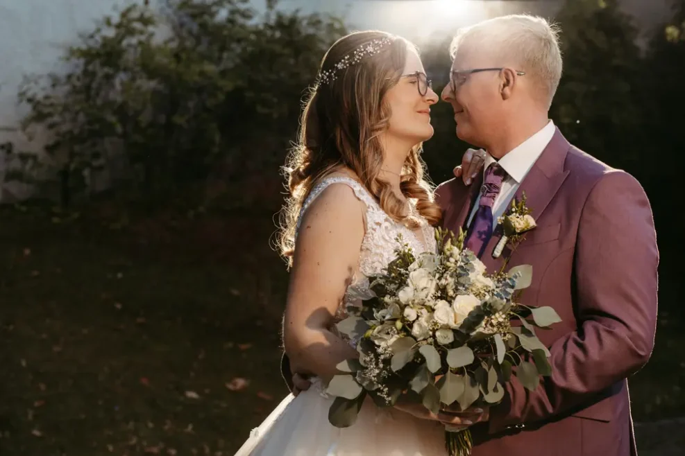 Eine Braut und ein Bräutigam stehen im Freien dicht beieinander und blicken sich in die Augen. Die Braut hält einen Strauß weißer Blumen und Grünzeug in der Hand, und das Sonnenlicht scheint hinter den beiden und schafft eine warme, romantische Atmosphäre.