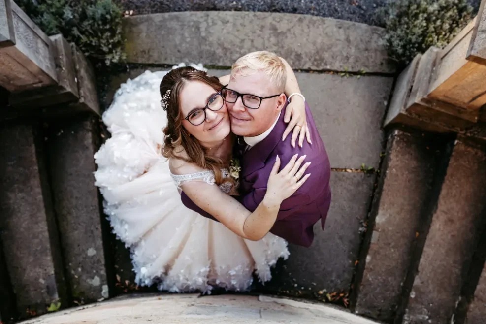 Eine Braut und ein Bräutigam mit Brille umarmen sich und lächeln in die Kamera. Die Braut trägt ein weißes Kleid und der Bräutigam einen violetten Anzug. Sie stehen auf steinernen Stufen, die von oben aufgenommen wurden.