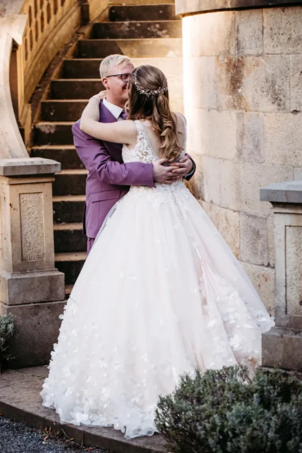 Eine Braut in einem weißen Kleid und ein Bräutigam in einem hellvioletten Anzug umarmen und küssen sich vor einer Steintreppe und einer rustikalen Steinmauer, im Freien bei natürlichem Licht.