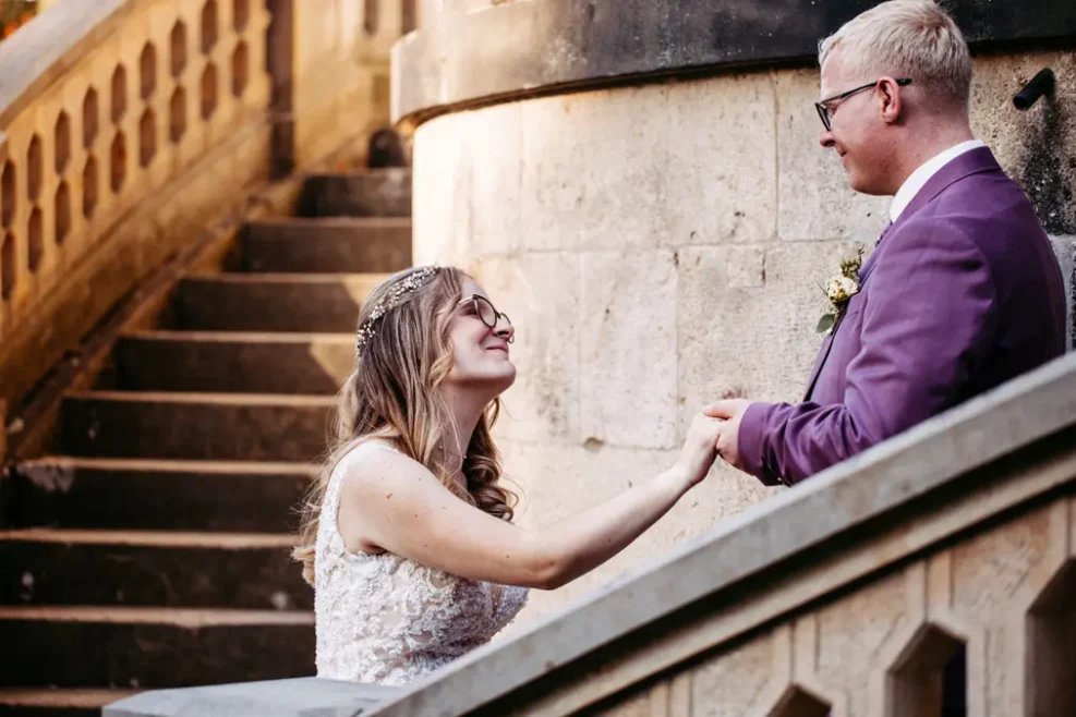 Eine Braut in einem weißen Kleid und ein Bräutigam in einem violetten Anzug halten sich an den Händen und lächeln sich auf den Steinstufen im Freien an, mit einer strukturierten Wand und Sonnenlicht im Hintergrund.
