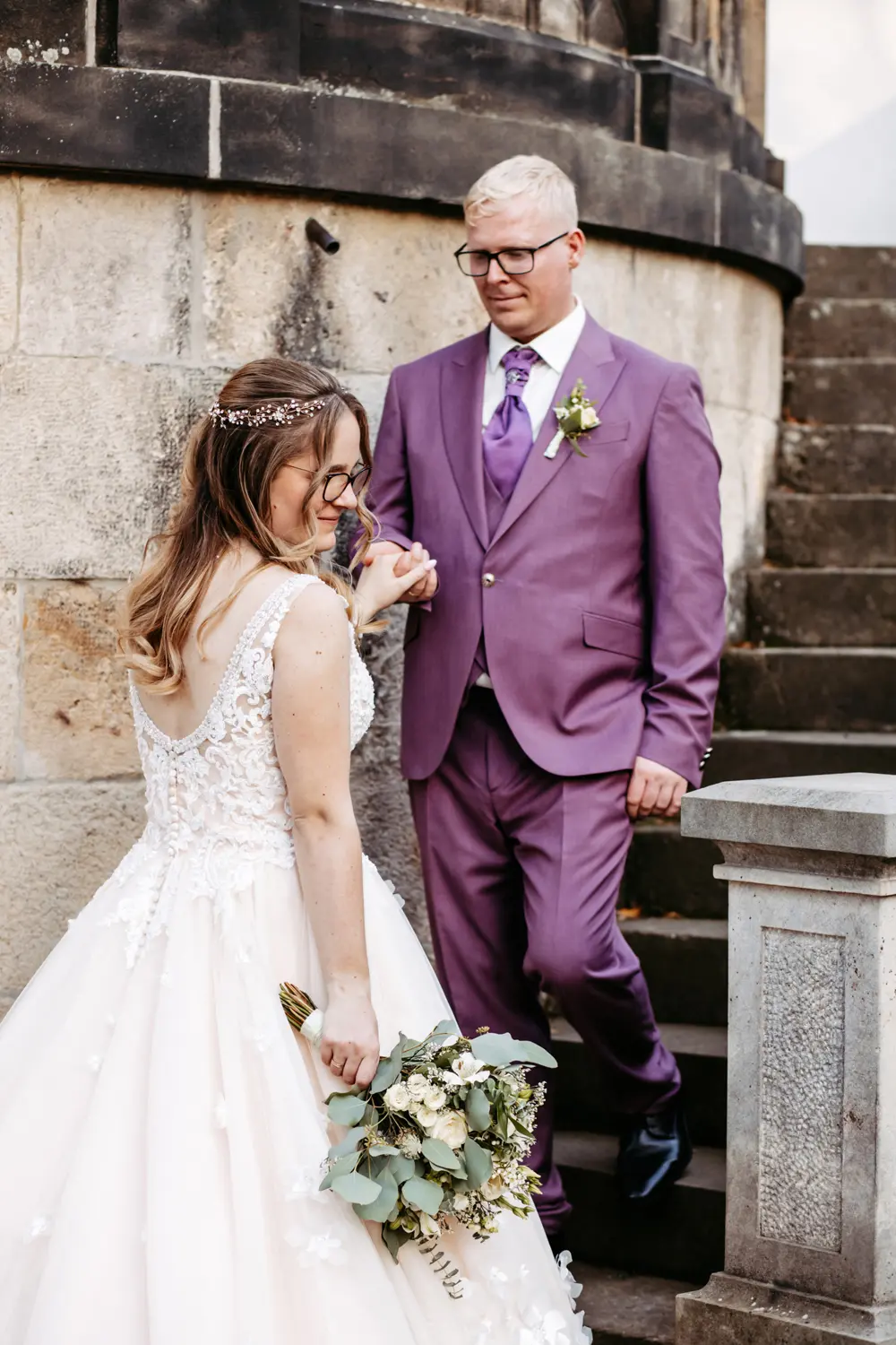 Eine Braut in einem weißen Spitzenkleid mit einem Blumenstrauß in der Hand wird von einem Bräutigam in einem violetten Anzug mit Krawatte die Steinstufen hinuntergeführt. Sie befinden sich im Freien in der Nähe eines Steingebäudes, und beide wirken fröhlich und elegant.