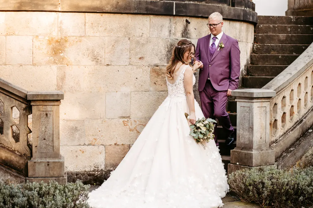Eine Braut in einem weißen Kleid und mit einem Blumenstrauß in der Hand steht am Fuße einer Steintreppe im Freien, während ein älterer Mann in einem violetten Anzug hinuntersteigt und sanft ihre Hand hält. Sie stehen neben einer Steinmauer mit Grünpflanzen in der Nähe.