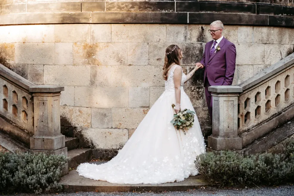 Eine Braut in einem weißen Kleid mit einem Blumenstrauß in der Hand steht auf steinernen Stufen und blickt zu einem Bräutigam in einem lila Anzug auf. Sie posieren vor einer verwitterten Steinmauer mit dekorativen Geländern auf beiden Seiten.