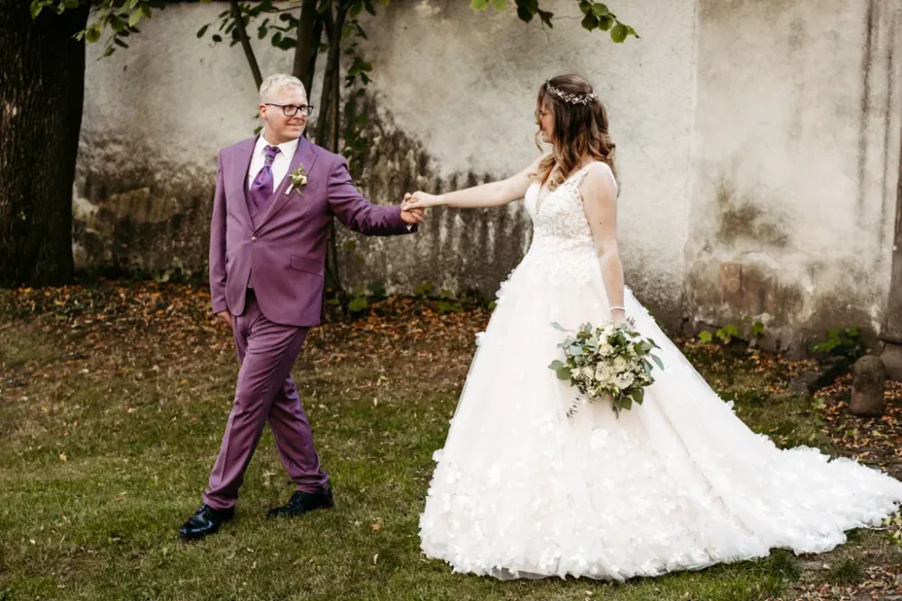 Eine Braut in einem weißen Kleid hält einen Blumenstrauß und die Hand des Bräutigams in einem lilafarbenen Anzug, während sie lächelnd über das Gras gehen, während im Hintergrund Bäume und eine strukturierte Wand zu sehen sind.