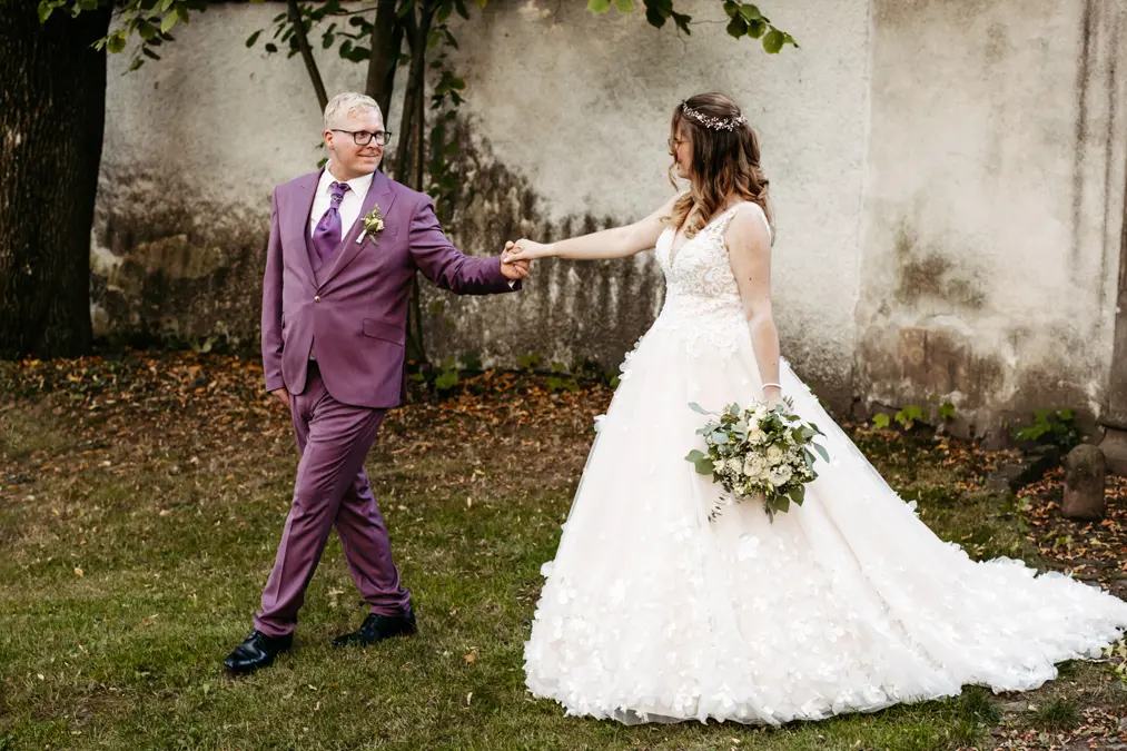 Eine Braut in einem weißen Kleid hält einen Blumenstrauß und die Hand des Bräutigams in einem lilafarbenen Anzug, während sie lächelnd über das Gras gehen, während im Hintergrund Bäume und eine strukturierte Wand zu sehen sind.