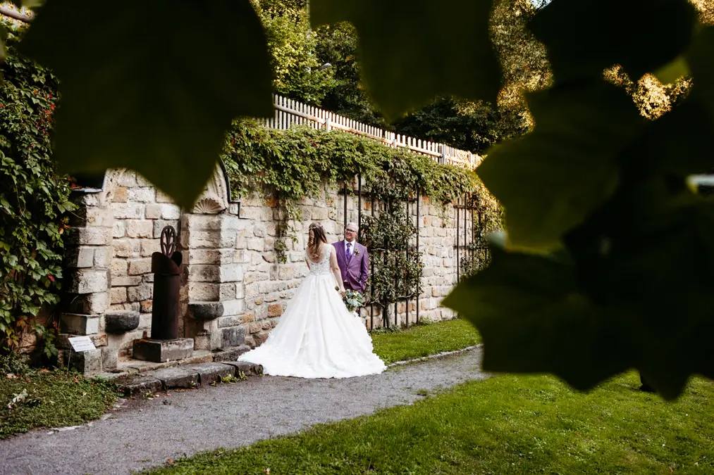 Eine Braut und ein Bräutigam stehen zusammen an einer Steinmauer, die mit Grün in einem Garten bedeckt ist, eingerahmt von großen, unscharfen Blättern im Vordergrund. Die Braut trägt ein weißes Kleid, und der Bräutigam trägt einen hellen Anzug.