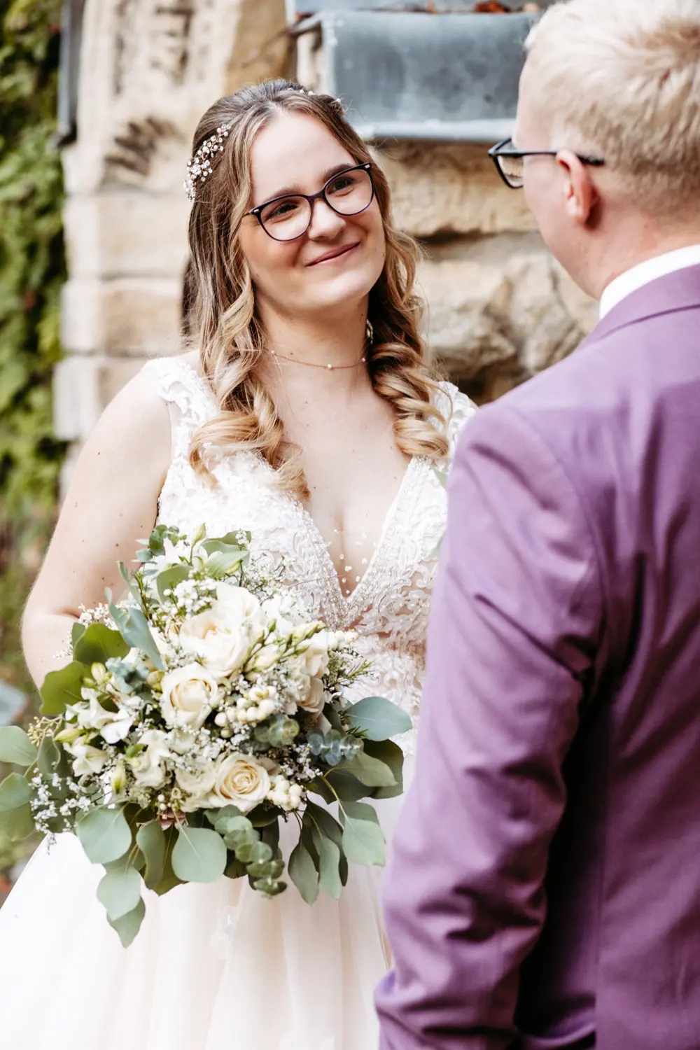 Eine Braut im weißen Kleid und mit Brille lächelt den Bräutigam im lavendelfarbenen Anzug liebevoll an, während sie einen Strauß weißer und grüner Blumen in der Hand hält, die im Freien an einer Steinmauer stehen.