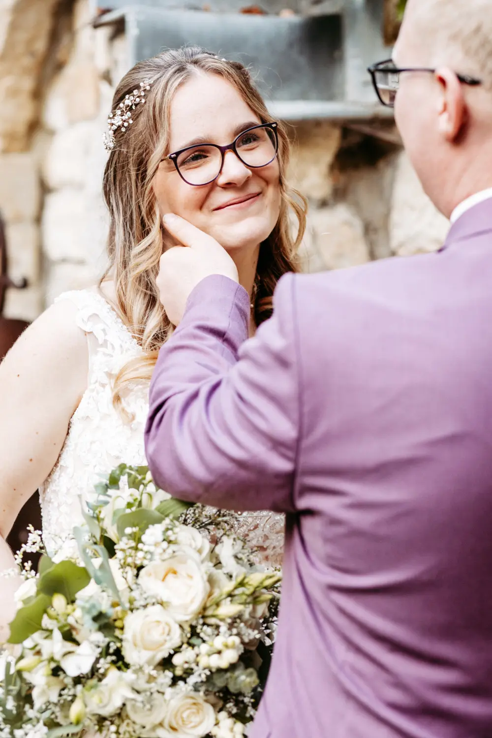 Eine Braut in einem weißen Kleid und mit Brille lächelt warm, als ihr Bräutigam in einem lavendelfarbenen Anzug sie sanft an der Wange berührt. Sie hält einen Strauß weißer und grüner Blumen in der Hand, und sie stehen an einer Steinmauer.