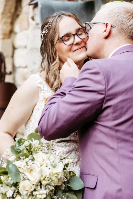 Ein Bräutigam in einem lavendelfarbenen Anzug küsst eine lächelnde Braut mit Brille und gewelltem Haar. Sie hält einen Strauß weißer Blumen und Grünzeug in der Hand. Sie stehen dicht beieinander vor einer Steinmauer.