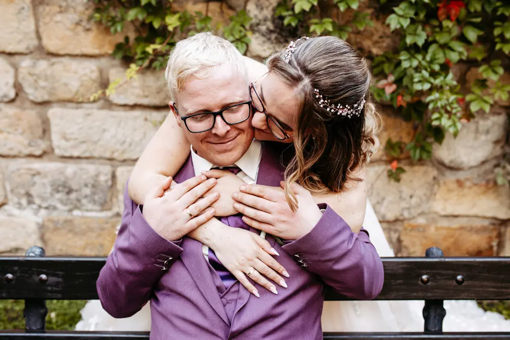 Eine Braut umarmt und küsst ihren lächelnden Bräutigam von hinten, während sie auf einer Bank sitzen. Beide tragen Brillen und formelle Kleidung, mit einer Steinmauer und Grünpflanzen im Hintergrund.