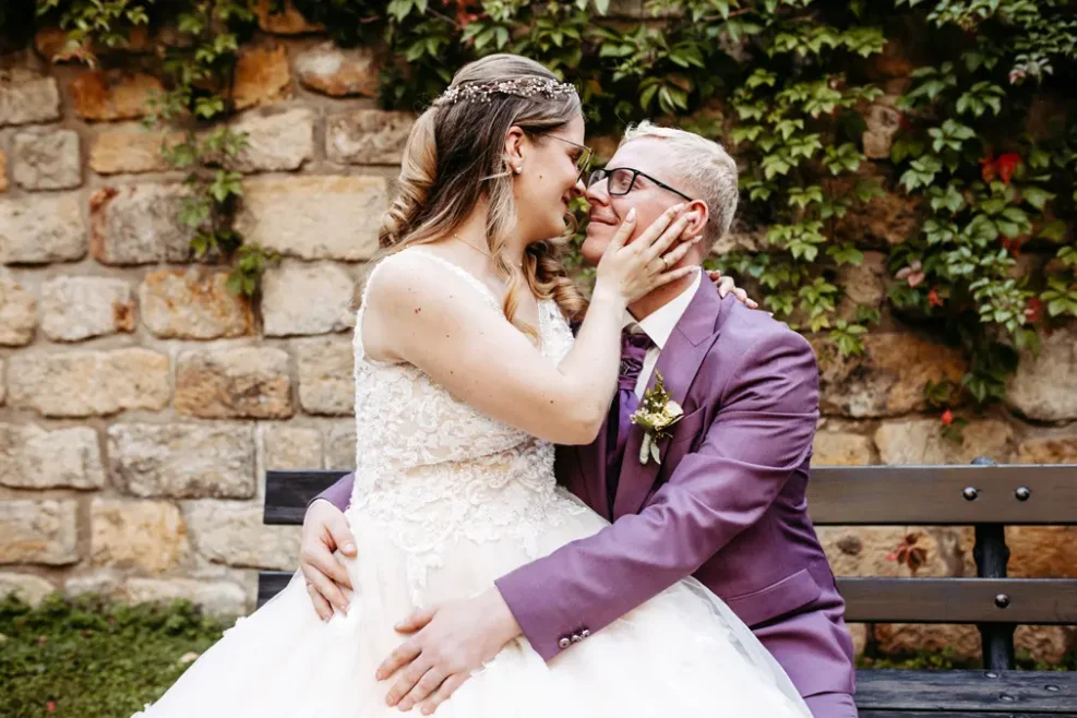 Ein frisch verheiratetes Paar sitzt auf einer Bank und blickt sich liebevoll in die Augen. Die Braut trägt ein weißes Spitzenkleid, der Bräutigam einen violetten Anzug. Sie sitzen dicht beieinander vor einer mit grünen Ranken bewachsenen Steinmauer.