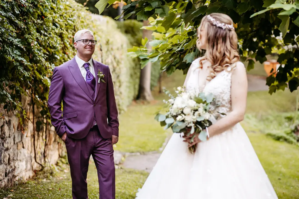 Ein Bräutigam in einem lila Anzug lächelt eine Braut in einem weißen Hochzeitskleid an, die einen Blumenstrauß hält. Sie stehen im Freien auf einem grasbewachsenen Weg in der Nähe einer Steinmauer und belaubter Bäume.