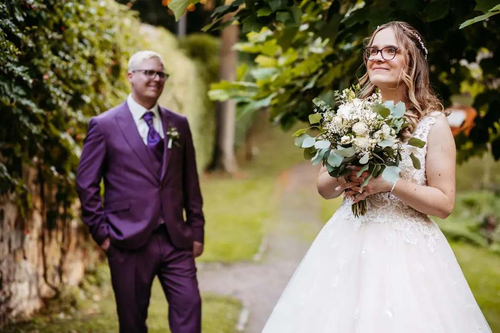 Eine Braut in einem weißen Kleid hält einen Blumenstrauß und lächelt, während der Bräutigam in einem lila Anzug hinter ihr steht und sie anschaut. Sie befinden sich im Freien auf einem von Bäumen gesäumten Weg.