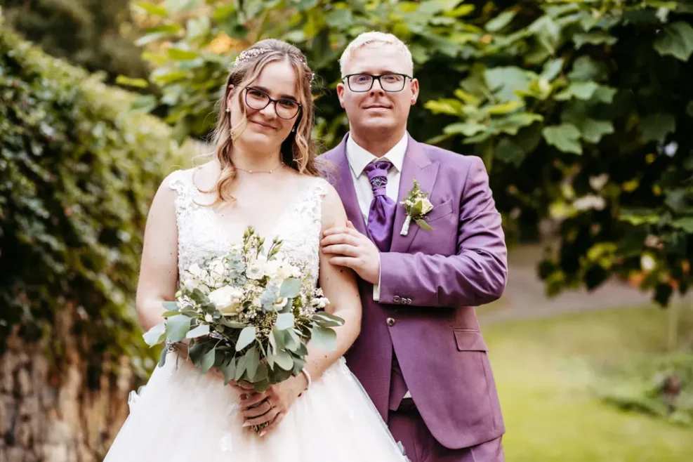 Eine Braut in einem weißen Kleid mit einem Blumenstrauß in der Hand steht neben einem Bräutigam in einem hellvioletten Anzug und einer Brille. Sie sind im Freien, umgeben von Grün, beide lächeln sanft in die Kamera.