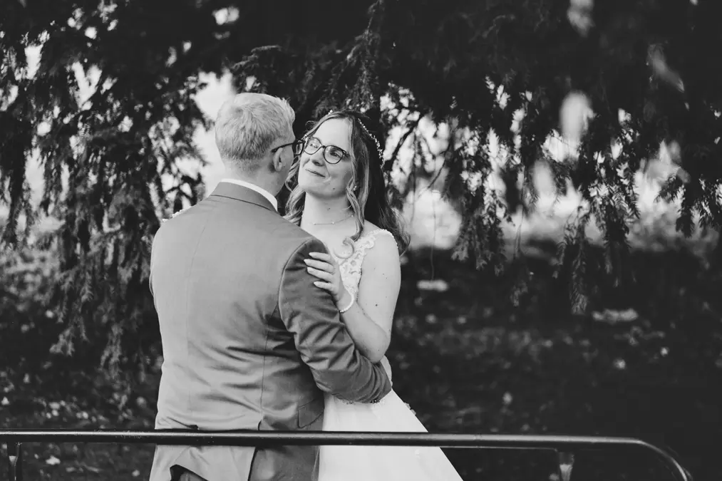 Ein Schwarz-Weiß-Foto eines Paares in Hochzeitskleidung, das sich im Freien umarmt, wobei die Braut lächelt und zum Bräutigam aufblickt, der unter den Zweigen eines Baumes steht.