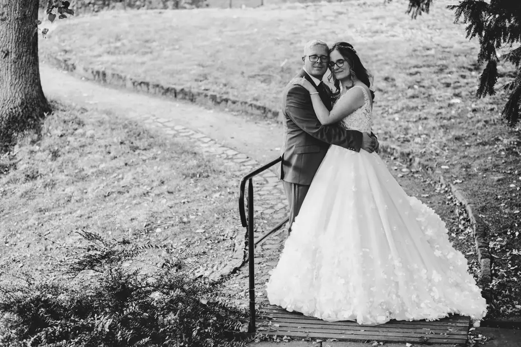 Eine Braut in einem weißen Kleid und ein Bräutigam im Anzug umarmen sich und lächeln auf einer kleinen Holzbrücke in einer parkähnlichen Umgebung, mit Bäumen und einem gewundenen Weg im Hintergrund. Das Foto ist in schwarz-weiß gehalten.