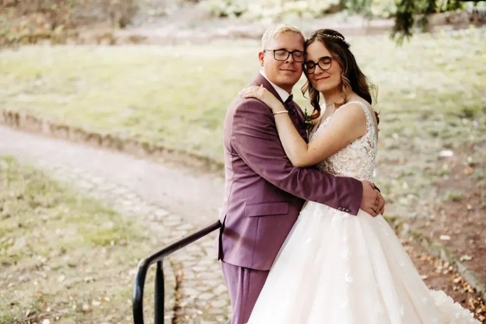 Eine Braut in einem weißen Hochzeitskleid und ein Bräutigam in einem lilafarbenen Anzug umarmen sich und lächeln im Freien auf einem von Grün umgebenen Steinweg.
